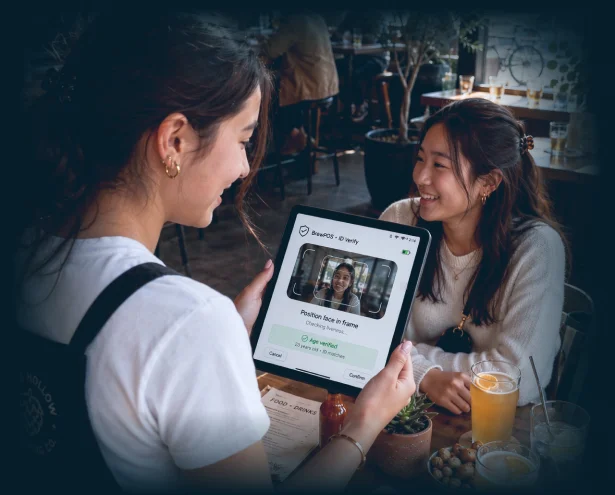 A server holding a tablet that shows a live face check and an age-verified result for a smiling guest at a restaurant table.