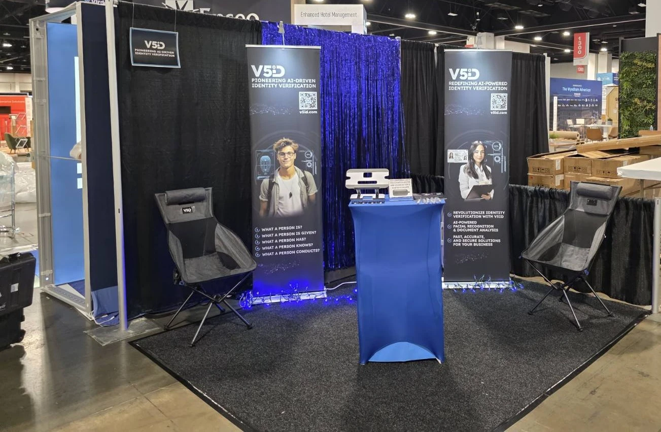 V5iD Booth Preparation: Setting the Stage for AI-Driven Identity Verification at The Hospitality Show 2025 in Denver, CO A vibrant trade show booth setup for V5iD during preparation for The Hospitality Show 2025, featuring sleek black and blue drapes, illuminated promotional banners highlighting "V5iD AI-Driven Identity Verification," a central blue pedestal table with demo equipment, ergonomic gray chairs, a verification kiosk with blue lighting, and QR code stands, all arranged on a gray carpeted floor amid the convention hall's ambient lighting and nearby exhibitor signage.