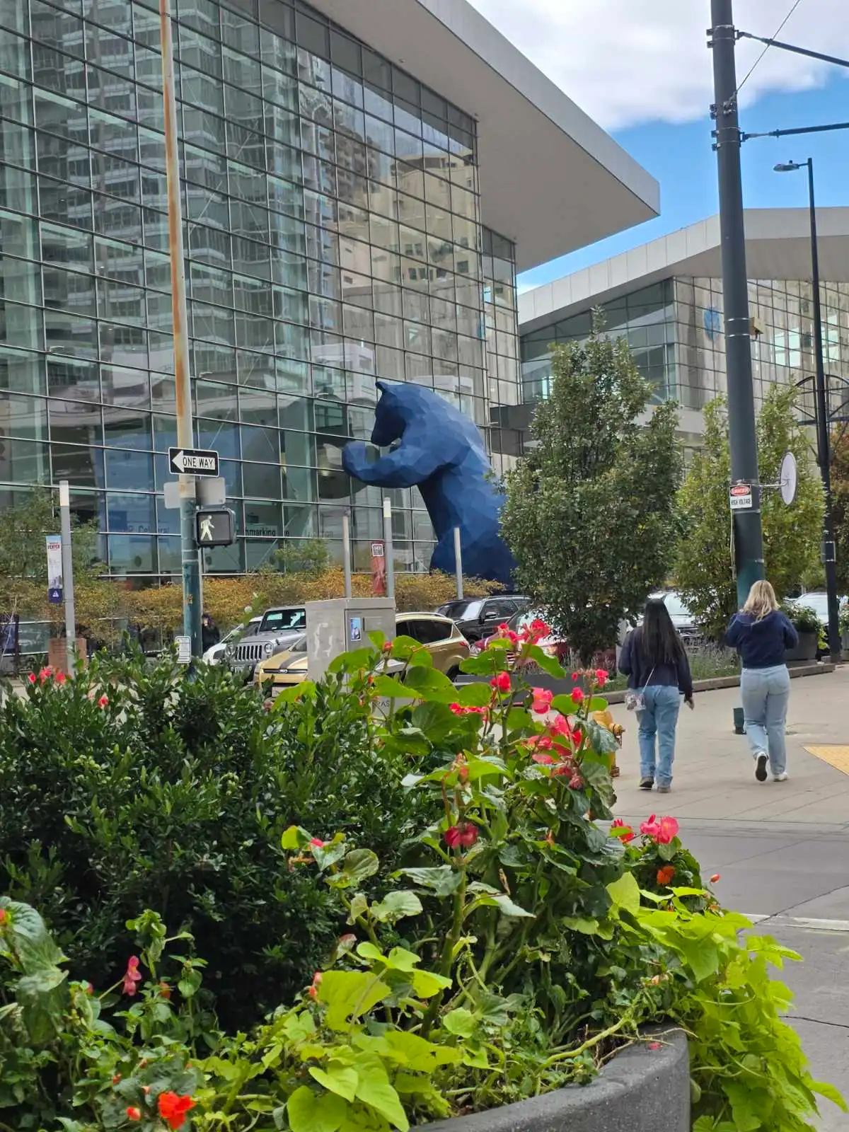 Approaching Innovation: The Iconic Colorado Convention Center at The Hospitality Show 2025 in Denver, CO A wide-angle view of the modern glass facade of the Colorado Convention Center in Denver during The Hospitality Show 2025, featuring the famous blue bear sculpture "I See What You Mean" playfully peeking into the building, surrounded by lush green shrubs and vibrant pink geranium flowers in concrete planters, with pedestrians in casual attire walking along the sidewalk near parked vehicles, under a partly cloudy sky with streetlights and directional signs adding to the bustling urban atmosphere.