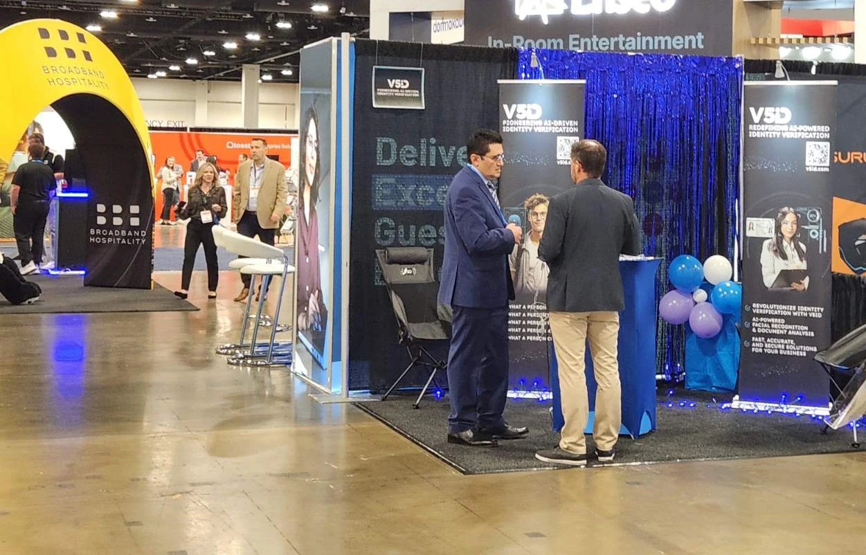 Engaging Conversations at the V5iD Booth: AI Identity Verification in Action at The Hospitality Show 2025 in Denver, CO V5iD booth at The Hospitality Show 2025, showing two suited professionals in discussion by a blue pedestal amid black drapes, "Deliver Exceptional Guest Experiences" banners, blue balloons, and demo chairs, with nearby BBB Hospitality arch and other exhibitors under convention hall lights.