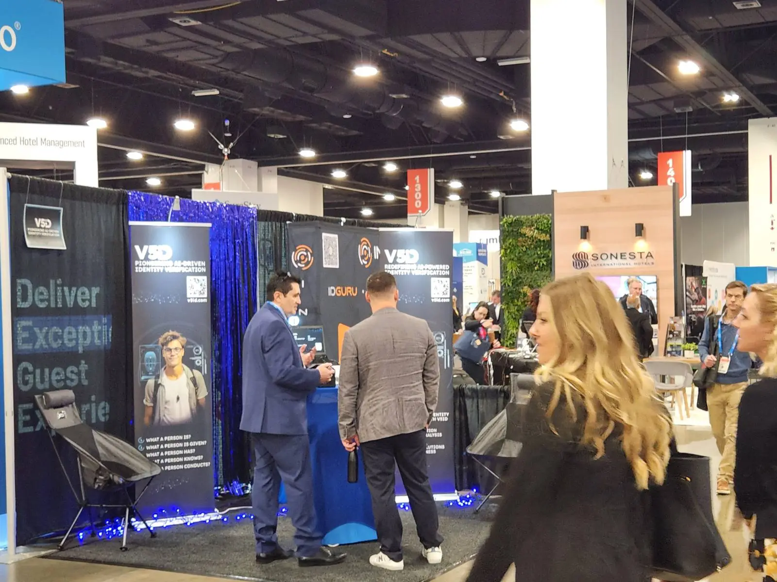 Live Demos and Networking: V5iD AI Verification Kiosk Draws Crowds at The Hospitality Show 2025 in Denver, CO V5iD booth at The Hospitality Show 2025, featuring a blue kiosk demo with suited professionals engaging a diverse group of attendees, promotional banners on identity verification, gray chairs, blue LED lights, and adjacent exhibitors like Sonesta with green wall under bright convention hall ceilings.