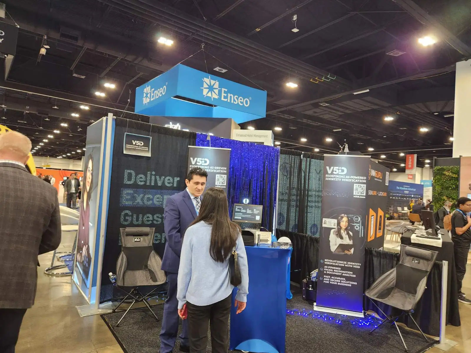Hands-On Demo: V5iD AI Identity Verification Captivates Attendees at The Hospitality Show 2025 in Denver, CO CTO of V5iD, demonstrating the AI-driven ID verification kiosk to an attendee at a pedestal with screens and scanners, surrounded by V5iD banners on pioneering identity verification, demo chairs, and LED accents in the convention hall.