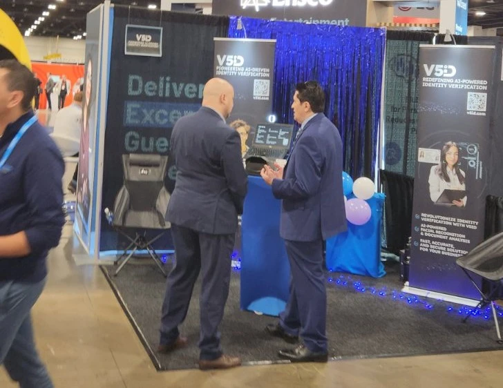 Dynamic Booth Interactions: V5iD Team Demonstrates AI Identity Verification at The Hospitality Show 2025 in Denver, CO V5iD booth at The Hospitality Show 2025, featuring two suited professionals—a bald man with beard and a dark-haired man—engaged in discussion by a blue pedestal with demo screens, "Deliver Exceptional Guest Experiences" banners, blue balloons, gray chair, and LED accents on a carpeted convention floor.