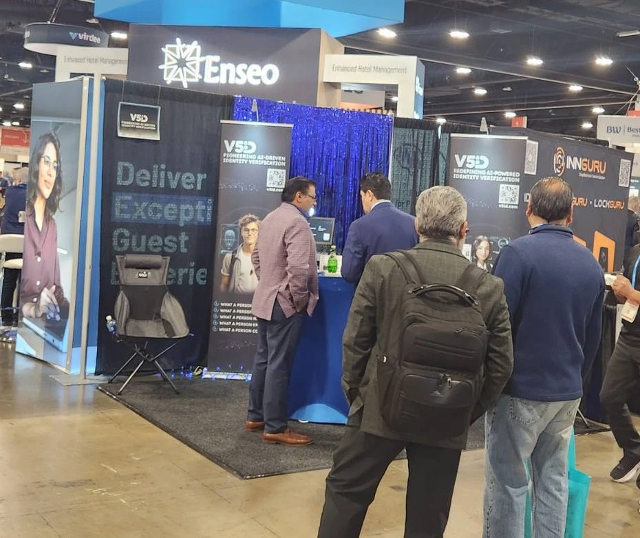 Professional Networking: V5iD Team Hosts Discussions on AI Identity Verification at The Hospitality Show 2025 in Denver, CO V5iD booth at The Hospitality Show 2025, featuring professionals engaging around a blue pedestal with verification monitors and scanners, V5iD banners on AI-driven identity solutions, demo chairs, blue drapes, and LED accents in the convention hall.