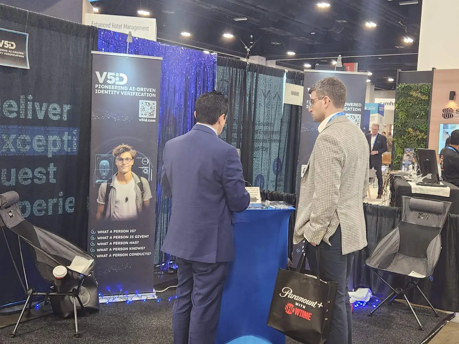 Collaborative Insights: V5iD Team Discusses AI Identity Solutions at The Hospitality Show 2025 in Denver, CO V5iD booth at The Hospitality Show 2025, showing professionals engaging at a blue pedestal with demo screens and scanners, banners highlighting pioneering AI-driven identity verification, gray chairs, blue drapes, and LED accents in the convention hall.
