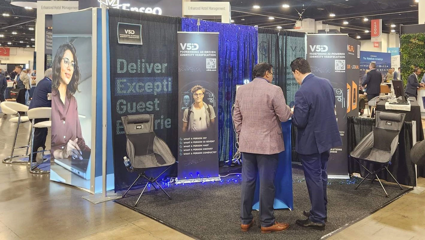 Team Engagement: V5iD Professionals Showcase AI Identity Verification at The Hospitality Show 2025 V5iD booth at The Hospitality Show 2025, featuring professionals discussing at a blue pedestal with verification displays, banners on pioneering AI-driven identity solutions, demo chairs, blue drapes, and LED accents in the convention hall.