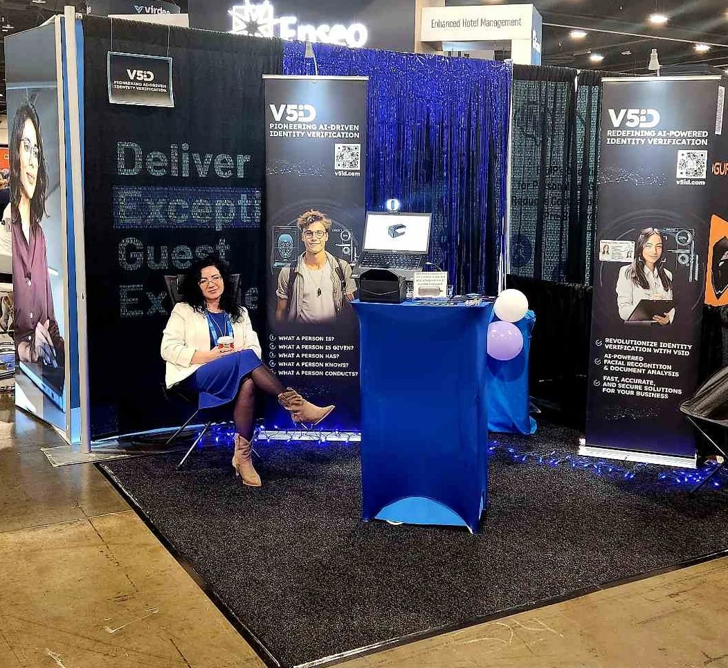 Inviting Discussions: V5iD Team Member Engages Visitors at The Hospitality Show 2025 in Denver, CO V5iD booth at The Hospitality Show 2025, showing a team member seated with a drink near a blue pedestal with verification screens and scanners, banners on pioneering AI-driven identity solutions, demo chairs, blue balloons, and LED accents in the convention hall.
