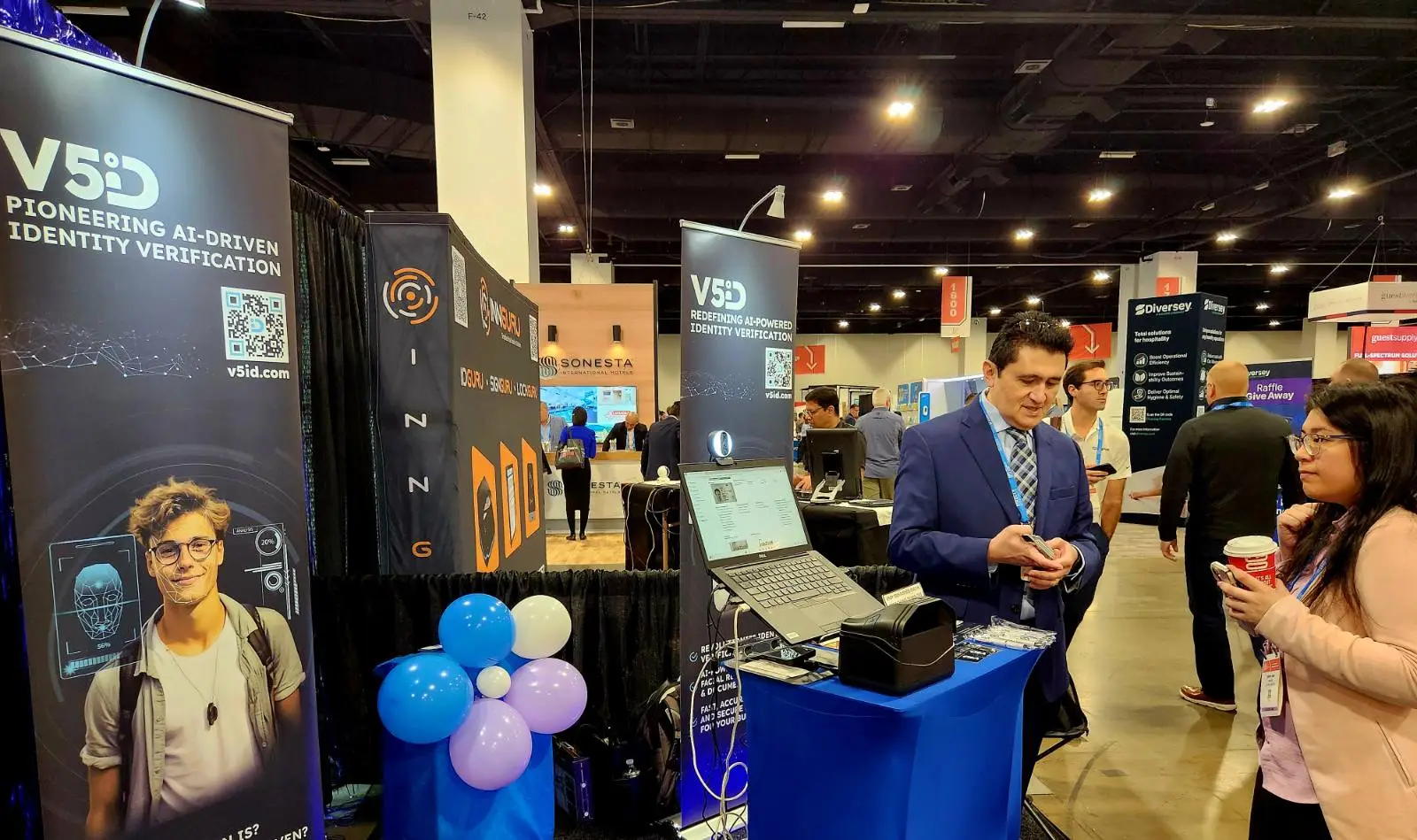 V5iD AI Identity Verification Demo: Engaging Attendees at The Hospitality Show 2025 Booth in Denver, CO V5iD booth demo at The Hospitality Show 2025, featuring a suited professional showcasing AI-driven identity verification kiosk with laptop and scanners to an attendee, surrounded by pioneering V5iD banners, QR codes, blue balloons, and LED accents on a convention floor.