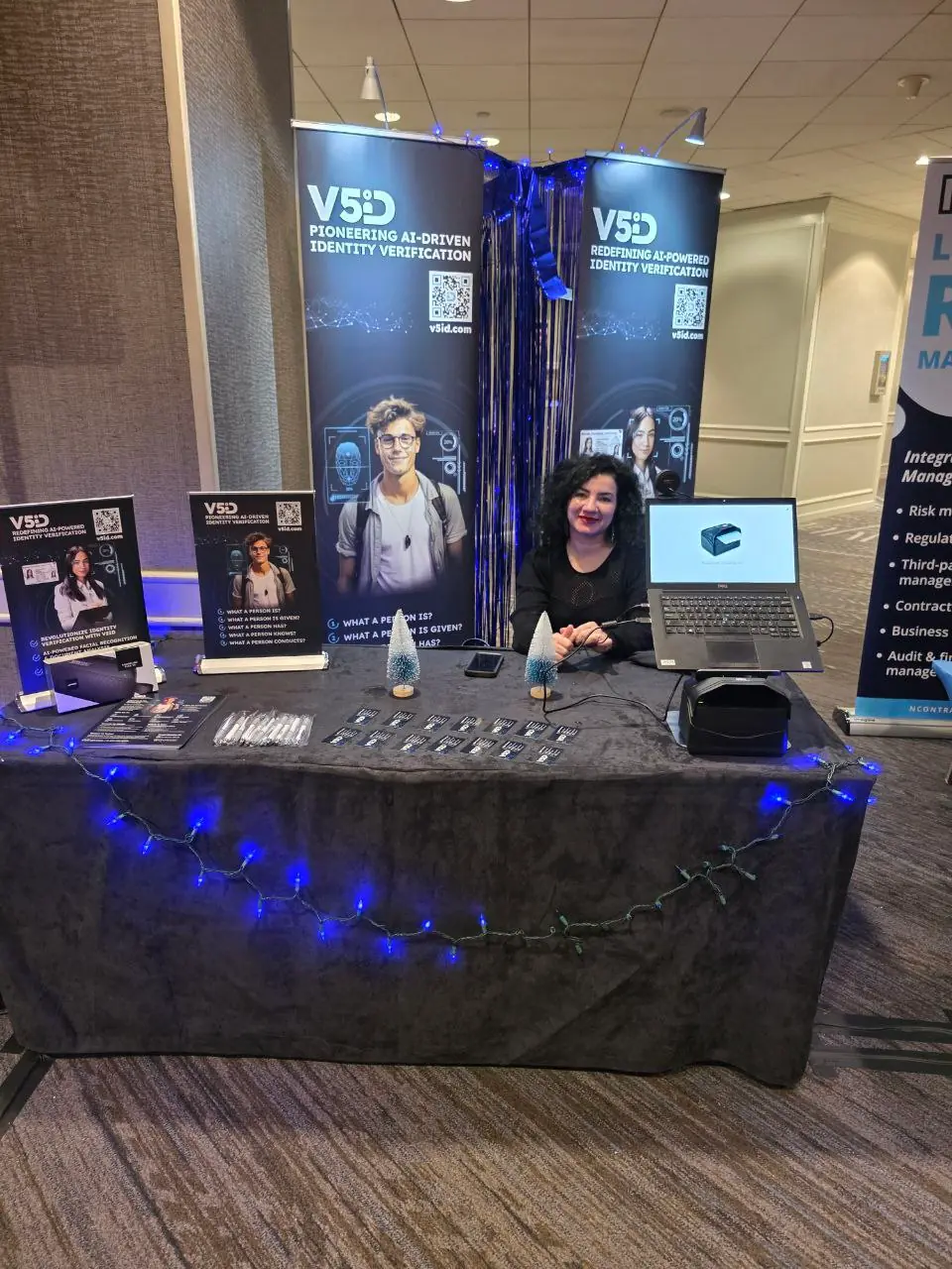 V5iD representative smiling at the company's booth during the Oregon Banking Leadership Symposium 2025 in Portland, featuring banners on pioneering AI-driven identity verification, QR codes to v5id.com, promotional materials, and blue holiday lighting