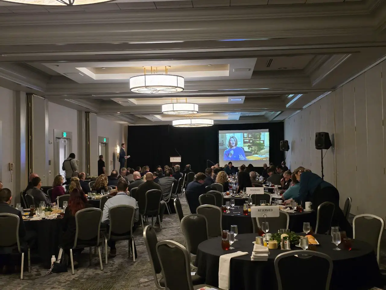 Banking professionals seated at round tables during a keynote presentation at the Oregon Banking Leadership Symposium 2025 in Portland, with a speaker at the podium and a large screen displaying a remote or featured speaker
