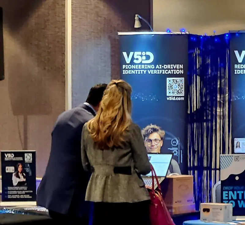 V5iD executive in suit and female attendee viewing a laptop demonstration at the company's booth during the Oregon Banking Leadership Symposium 2025 in Portland, with banners on pioneering AI-driven identity verification, QR codes to v5id.com, blue lighting