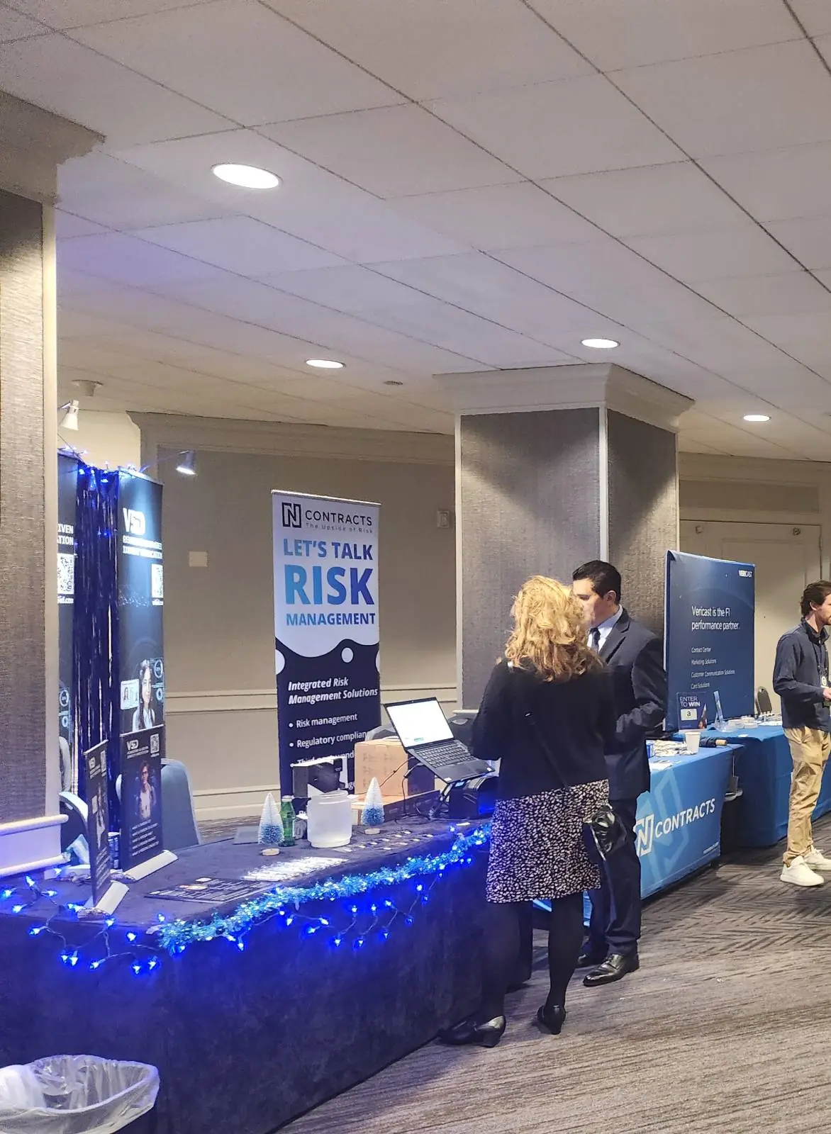 V5iD executive in suit engaging in conversation with a female attendee at the company's AI-driven identity verification booth during the Oregon Banking Leadership Symposium 2025 in Portland, featuring blue lighting, promotional banners with QR codes to v5id.com, laptop display