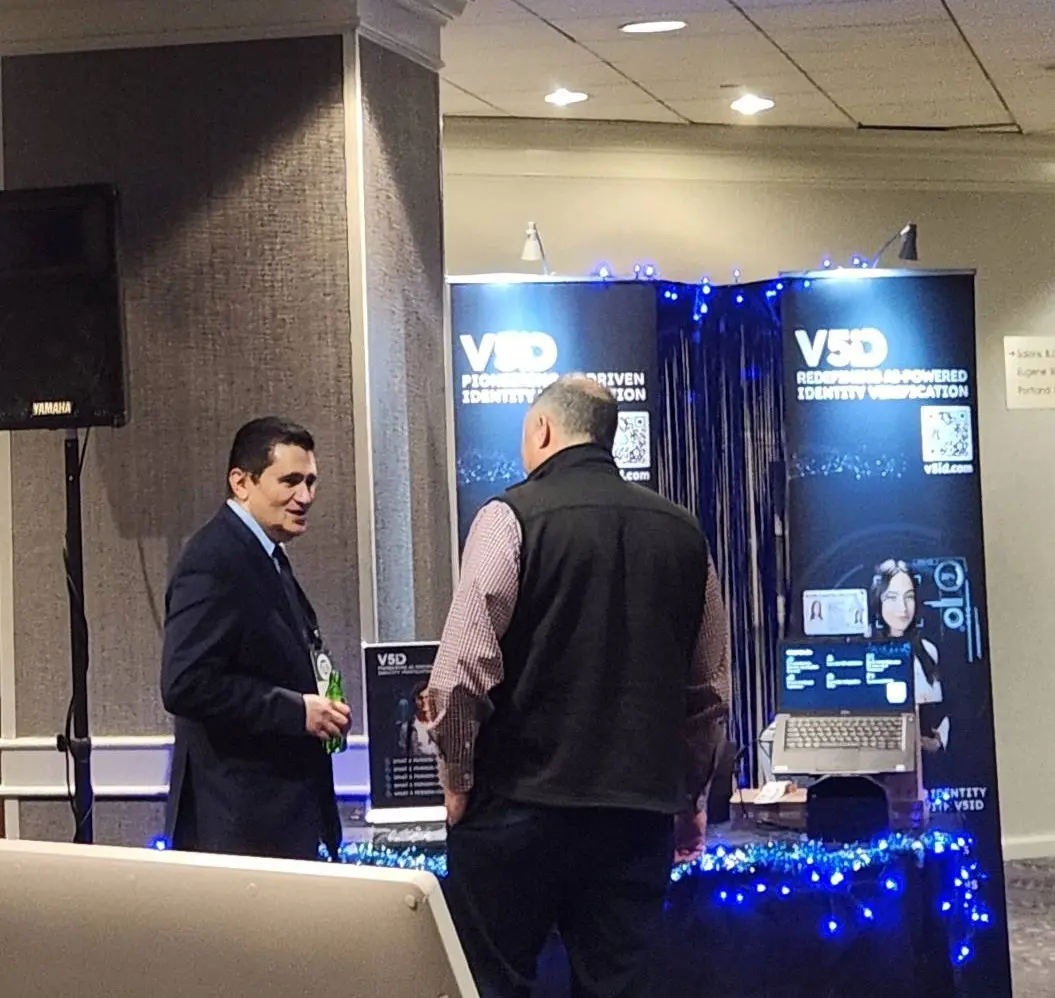 V5iD executive in suit holding a drink while conversing with an attendee at the company's booth during the Oregon Banking Leadership Symposium 2025 in Portland, featuring banners on pioneering and redefining AI-driven identity verification, QR codes to v5id.com, laptop display, promotional materials, and blue decorative lighting