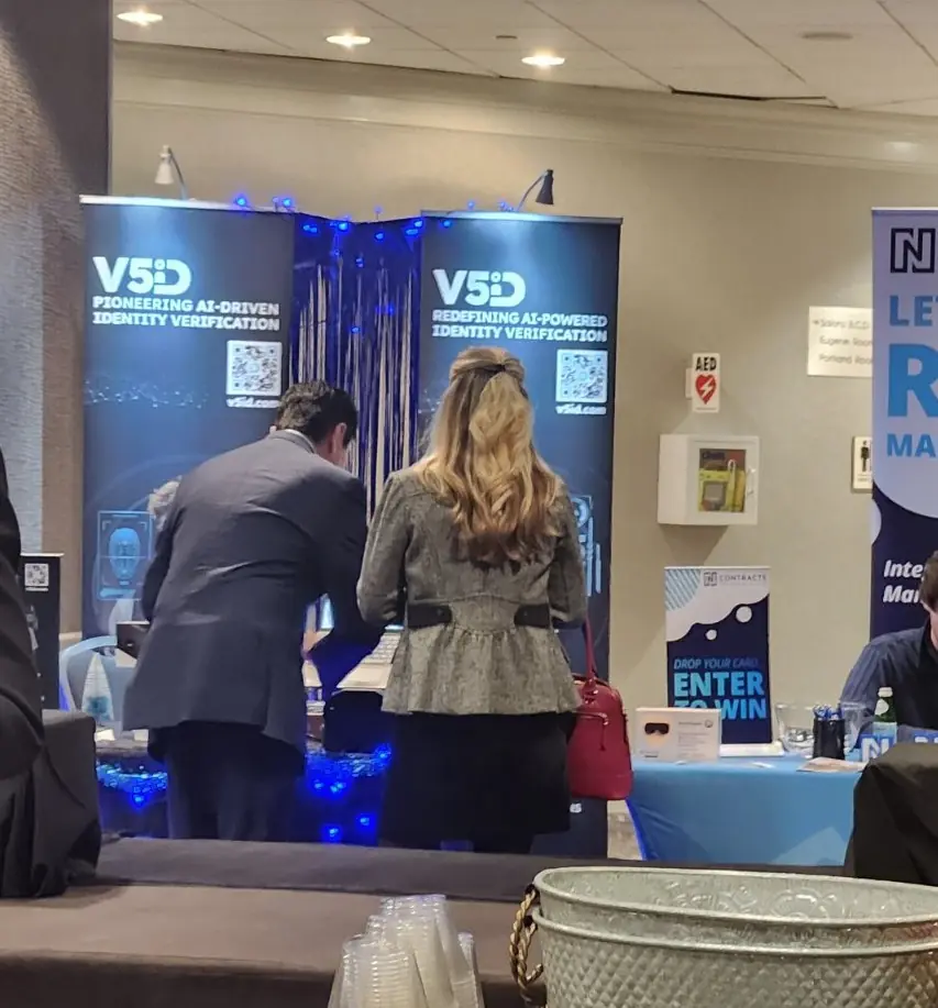 Attendees, including an executive in suit and a woman with blonde hair, engaging with a demonstration or materials at the V5iD booth during the Oregon Banking Leadership Symposium 2025 in Portland, featuring banners on pioneering and redefining AI-powered identity verification, QR codes to v5id.com, blue decorative lighting