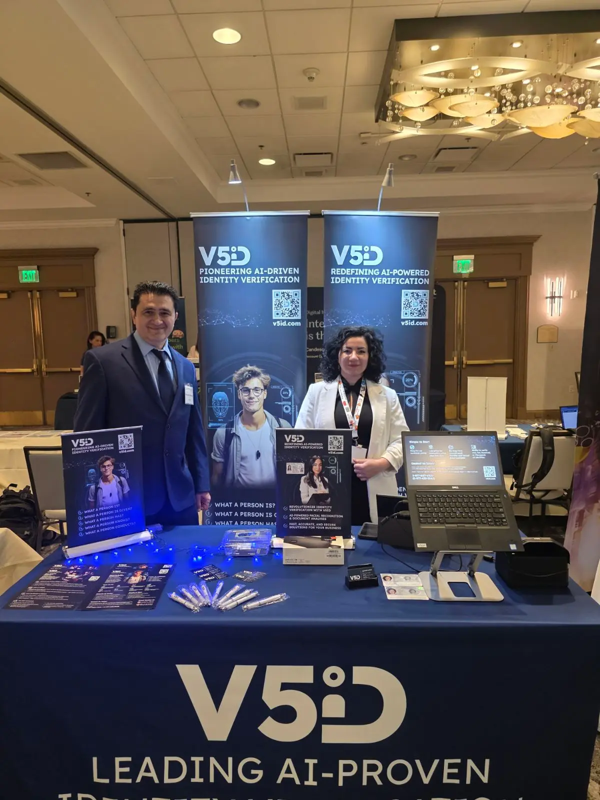 V5iD® CTO and CEO standing proudly behind their branded conference booth at SymWest 2026, surrounded by AI identity verification banners and demo equipment