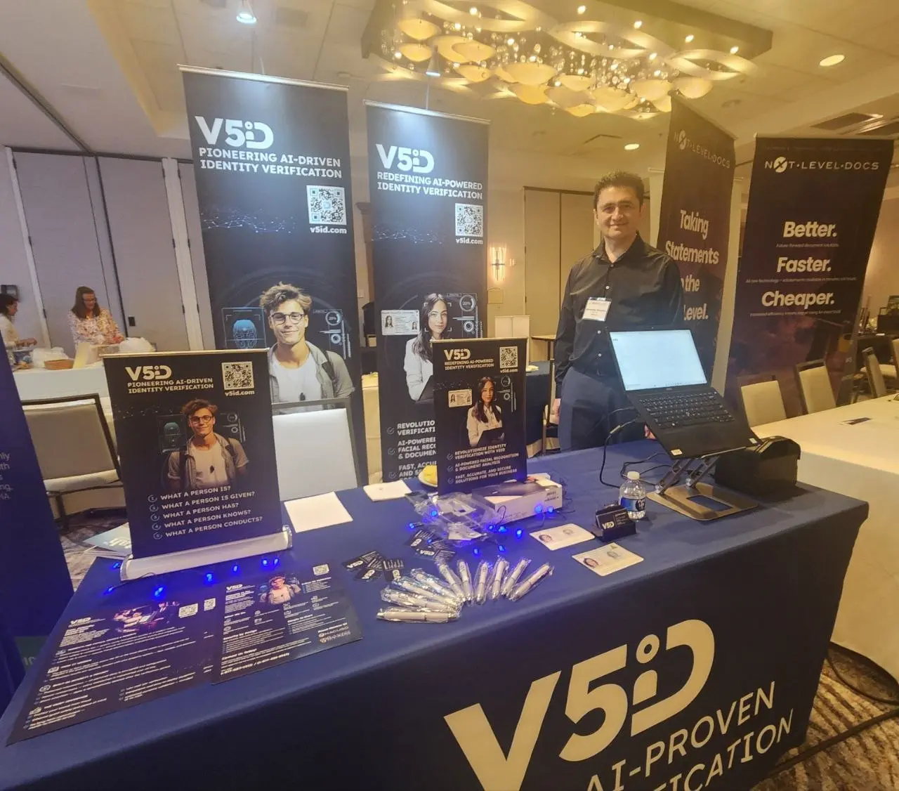 V5iD CTO standing behind the company's branded exhibit booth on day two of the SymWest conference, with AI identity verification banners, promotional materials, and demo laptops displayed