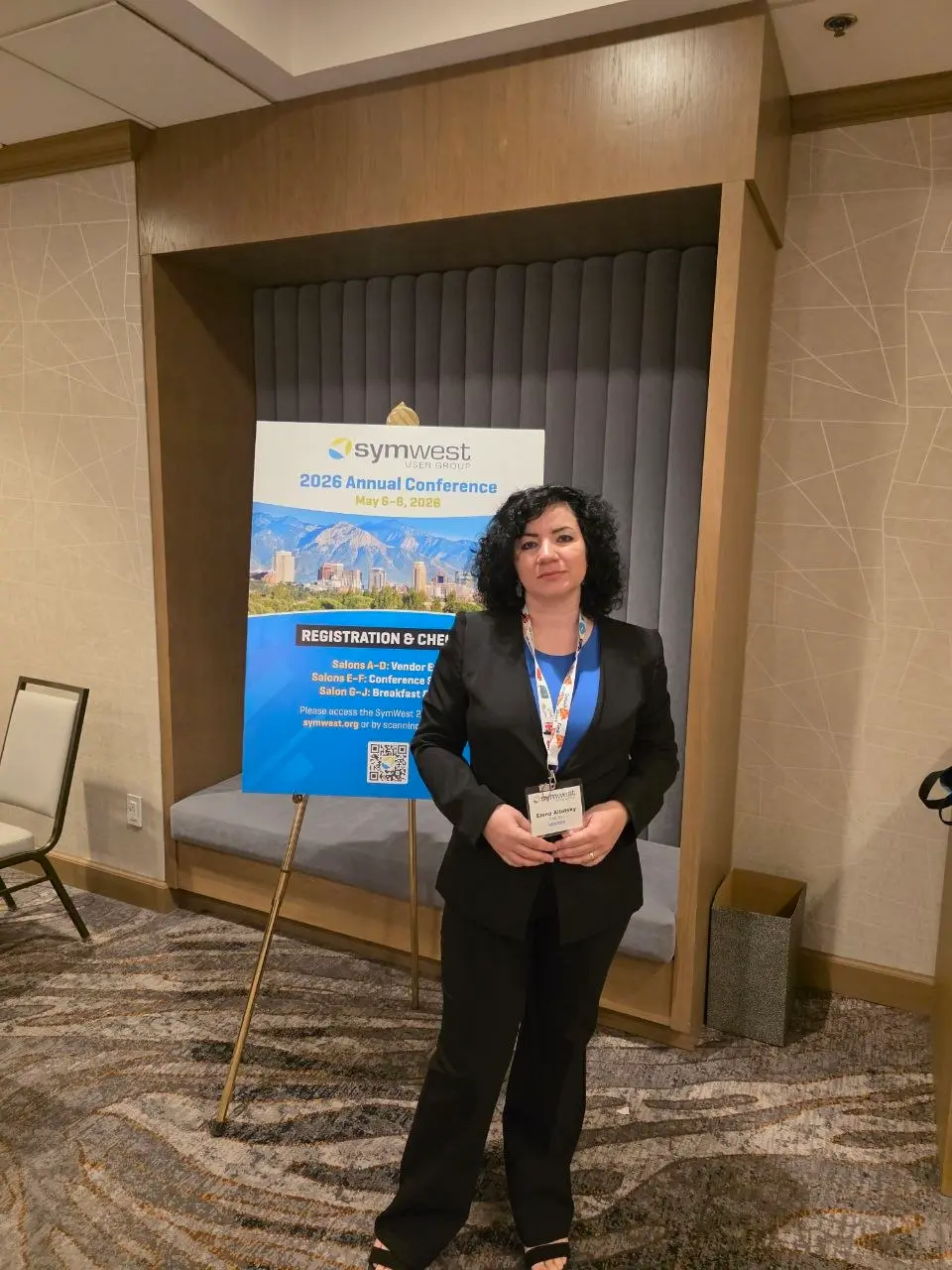 V5iD CEO standing in front of the SymWest 2026 Annual Conference registration sign at the Salt Lake Marriott Downtown, wearing her vendor badge
