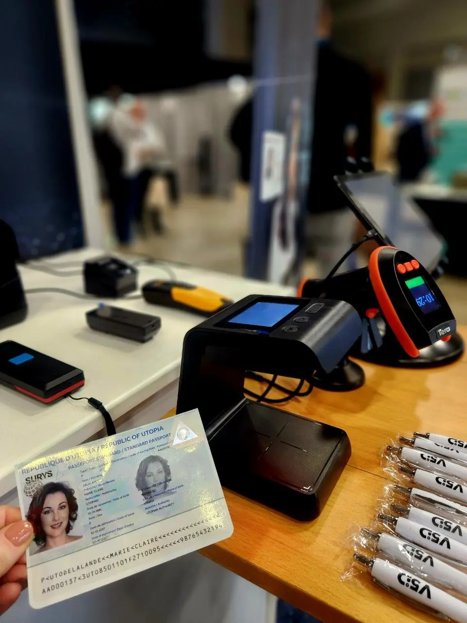 Close-up of V5iD identity verification demo showing a sample passport held next to biometric scanners, document readers and GSA-branded pens at Identity Week America 2026
