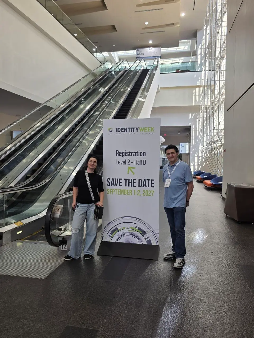 Two V5iD representatives standing beside the Identity Week America registration directional sign near escalators at the Walter E. Washington Convention Center
